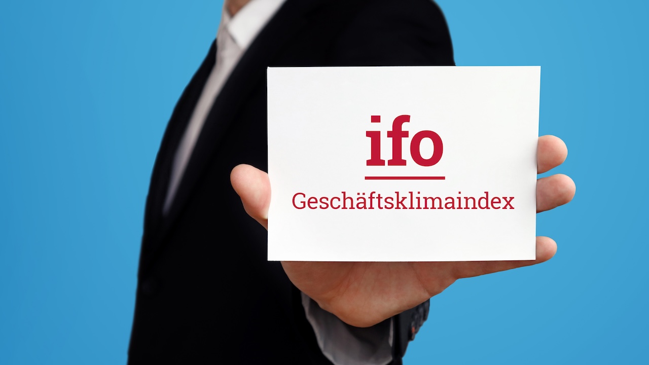 Geschäftsmann im Anzug hält eine Karte mit dem Schriftzug „ifo Geschäftsklimaindex“ vor blauem Hintergrund