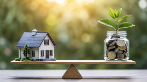 Modellhaus und mit Münzen gefülltes Sparglas mit Pflanze liegen auf einer Waage im Gleichgewicht.
