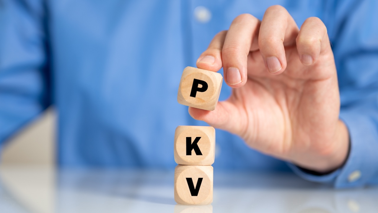 Hand stapelt drei Holzwürfel mit den Buchstaben P, K und V vor blauem Hintergrund.