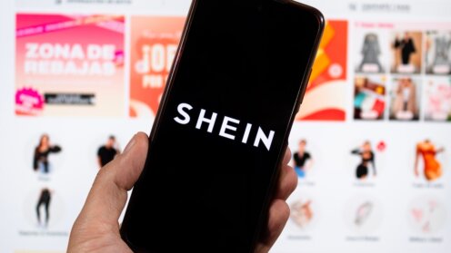 Eine Hand hält ein Smartphone mit dem SHEIN-Logo vor einer unscharf dargestellten Shopping-Webseite.