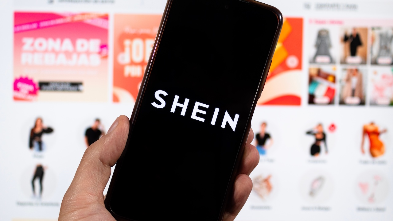 Eine Hand hält ein Smartphone mit dem SHEIN-Logo vor einer unscharf dargestellten Shopping-Webseite.