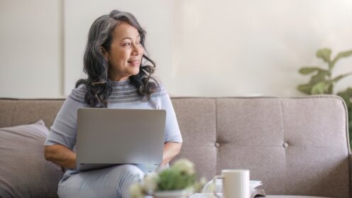 Frau mittleren Alters sitz mit Laptop auf dem Schoß auf der Couch.