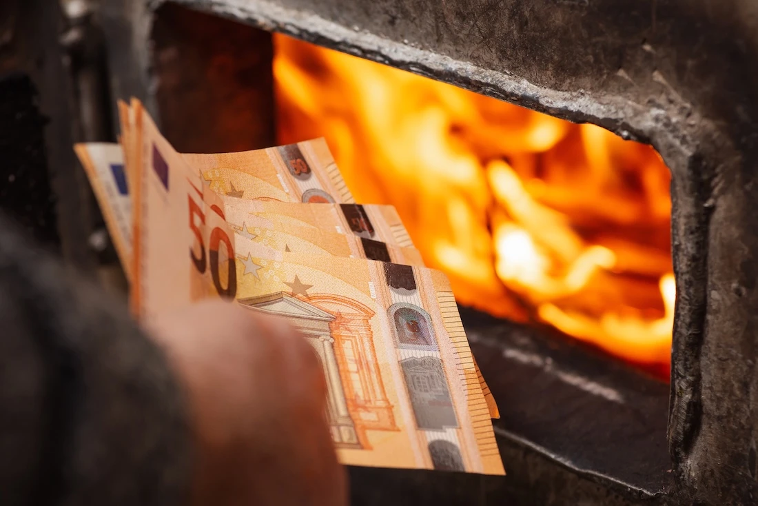 Euro-Scheine in der Hand eines Mannes neben dem Heizkessel, dessen Türen offen stehen und aus dem Flammen lodern. Hohe Ausgaben für die Beheizung des Hauses, teure Heizkosten