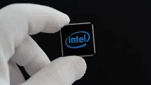 Eine behandschuhte Hand hält einen kleinen schwarzen Mikrochip mit dem blauen Intel-Logo vor schwarzem Hintergrund.